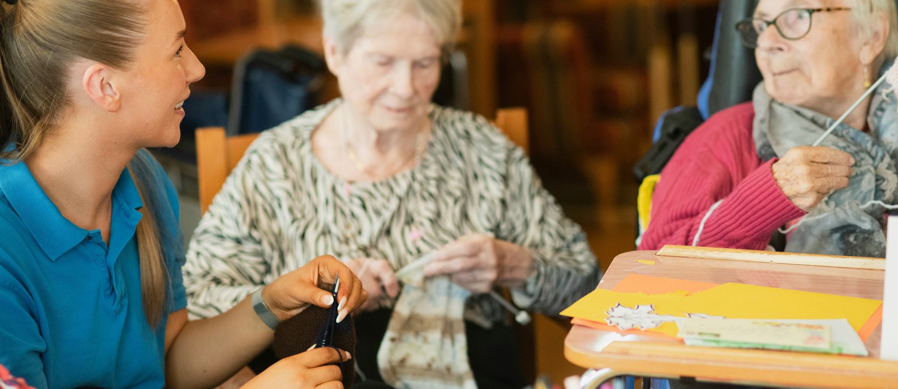 A carer sitting with two residents at a craft table, knitting and talking.