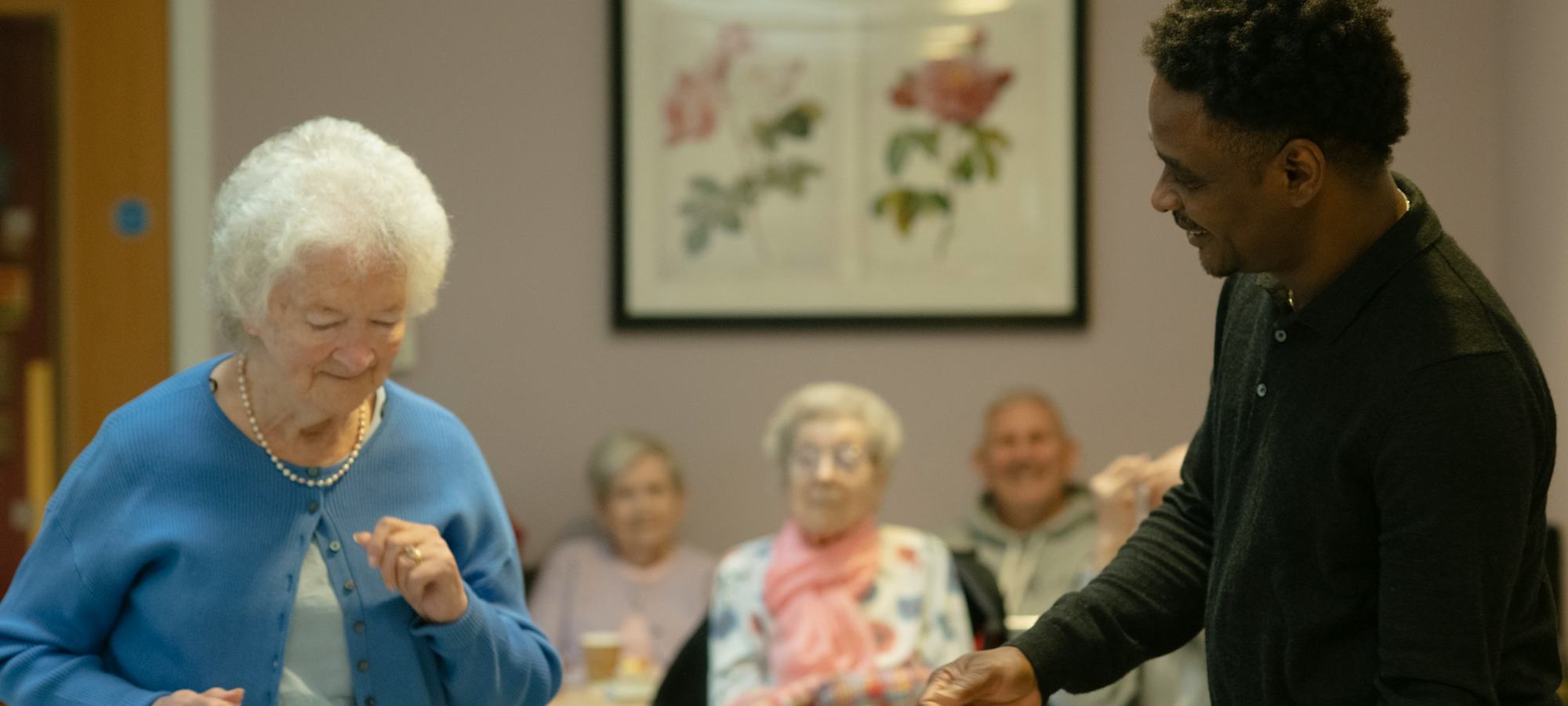 A member of staff dancing with a resident in a care home lounge while others watch and smile.