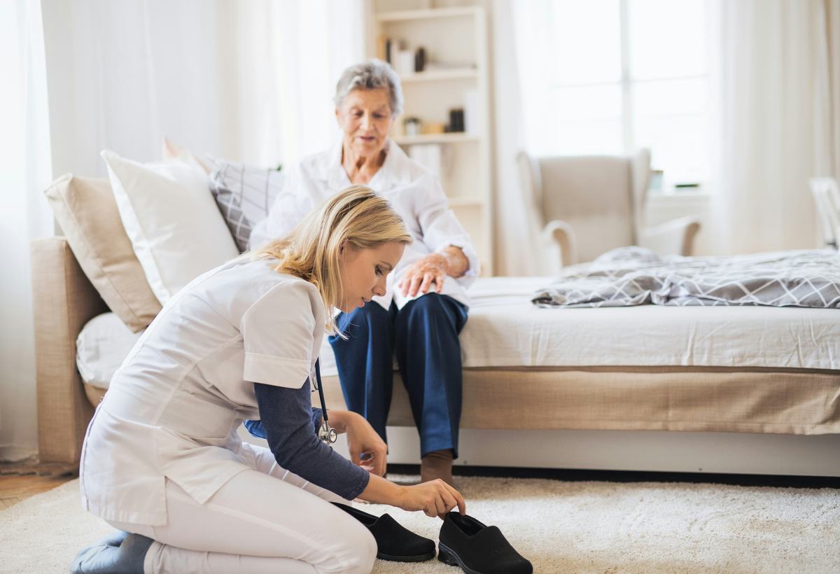 A carer kneeling to help a resident put her shoes on at the side of the bed.